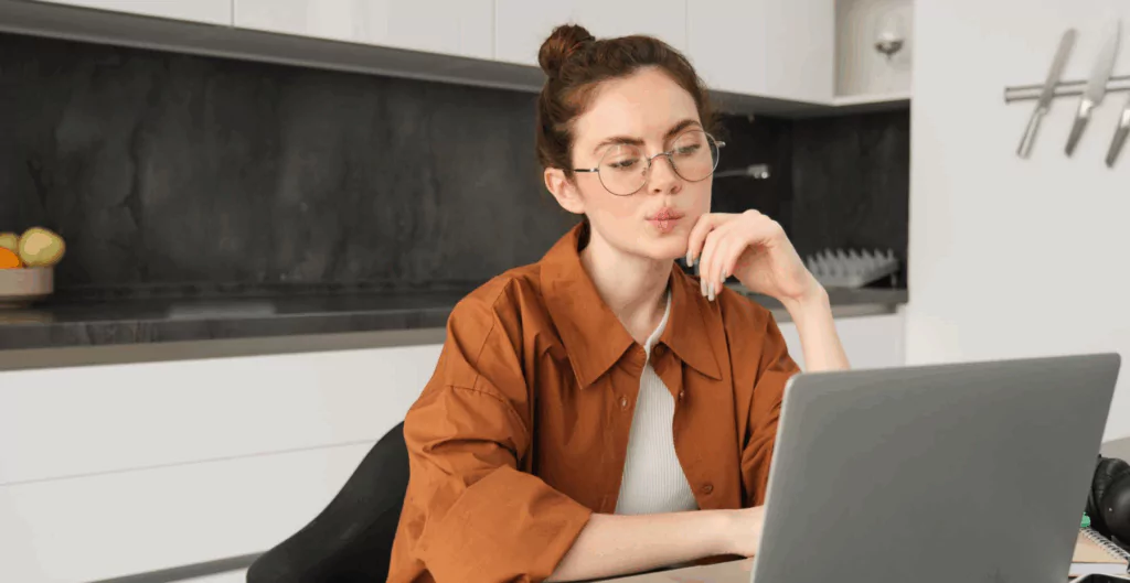 Mulher jovem sentada em mesa olhando para notebook com cozinha ao fundo