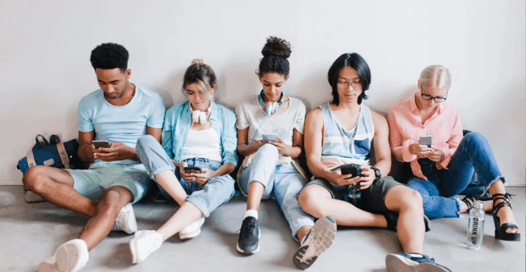 cinco jovens diferentes, sentados no chão, mexendo no celular, com roupas esportivas
