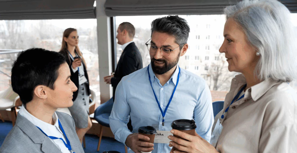 Homens e mulheres profissionais conversando durante um evento de networking, segurando copos de café, com cenários urbanos ao fundo.
