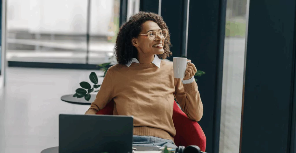 Mulher segurando caneca, sorrindo com escritório ao fundo 