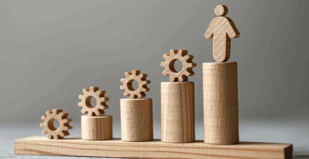 Imagem com blocos de madeira formando uma escada ascendente, com engrenagens e uma figura de pessoa no topo, representando crescimento e desenvolvimento pessoal ou profissional.