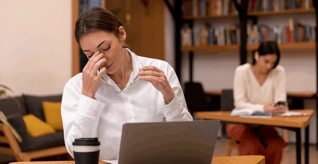 Mulher jovem com expressão de estresse e cansaço trabalhando em um ambiente de escritório, com outra pessoa ao fundo usando celular.