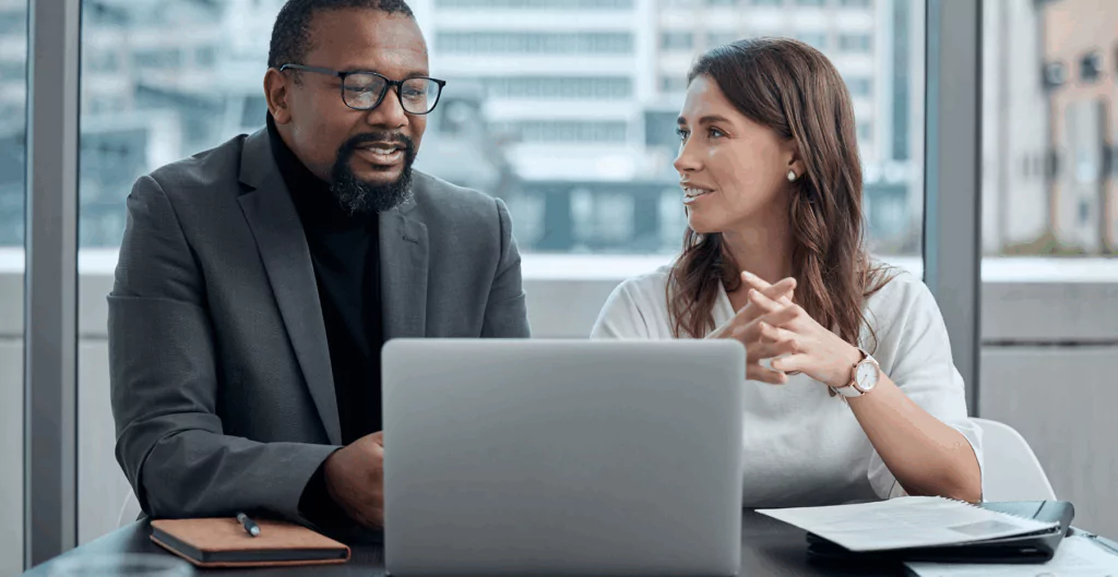 Duas pessoas conversando na frente de um notebook e uma mesa de escritório