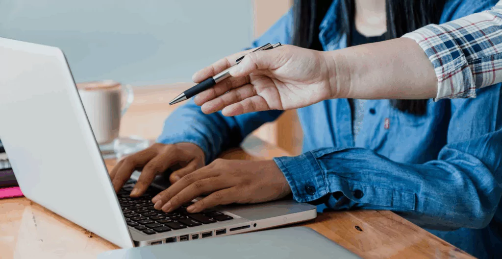 Pessoa digitando em notebook e uma outra pessoa apontando a tela com a caneta 
