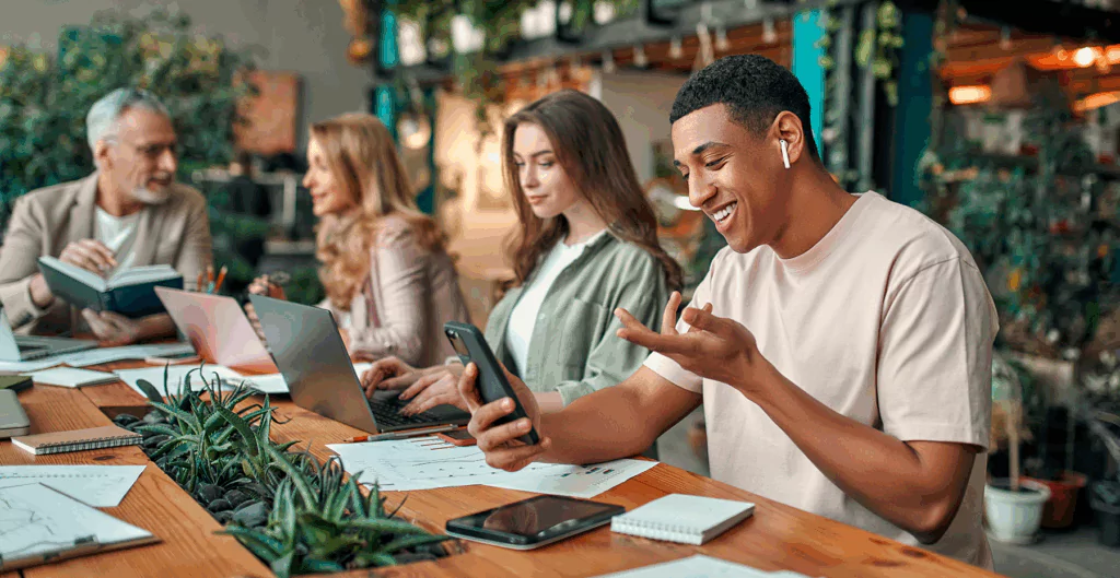 Jovem usando fones de ouvido e smartphone em ambiente de coworking, rodeado por colegas de trabalho e notebooks, em um espaço moderno e acolhedor.