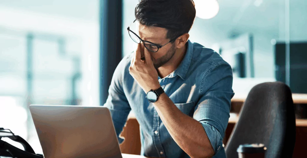 Homem jovem de óculos com expressão de preocupação enquanto trabalha em um laptop em um ambiente de escritório moderno.