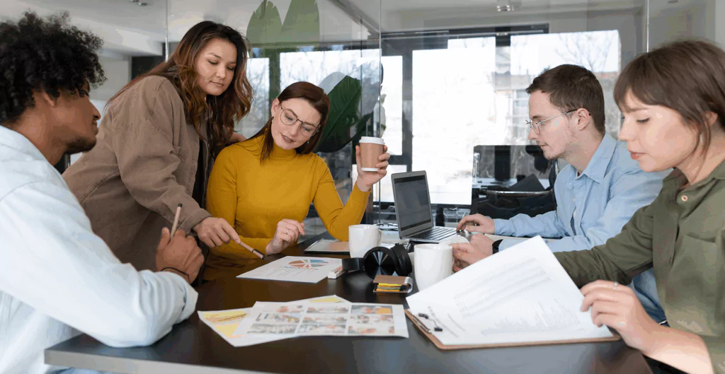 Equipe de profissionais de negócios colaborando em reunião de trabalho, discutindo projetos e estratégias, com laptops, papéis e canetas em uma sala de escritório moderna.