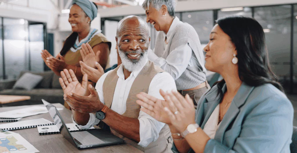 Imagem de um grupo de pessoas diversas comemorando e sorrindo em um ambiente de escritório, indicando inclusão e trabalho em equipe.