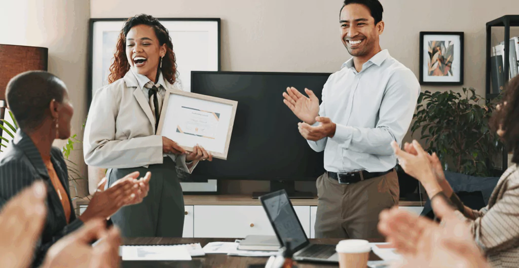 Mulher segurando certificado de sucessão de liderança com outras pessoas ao lado aplaudindo, com escritório ao fundo