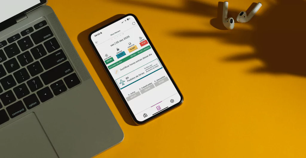 Celular mostrando aplicativo de controle de ponto da Pontotel, acompanhado de laptop e fones de ouvido, sobre mesa amarela.