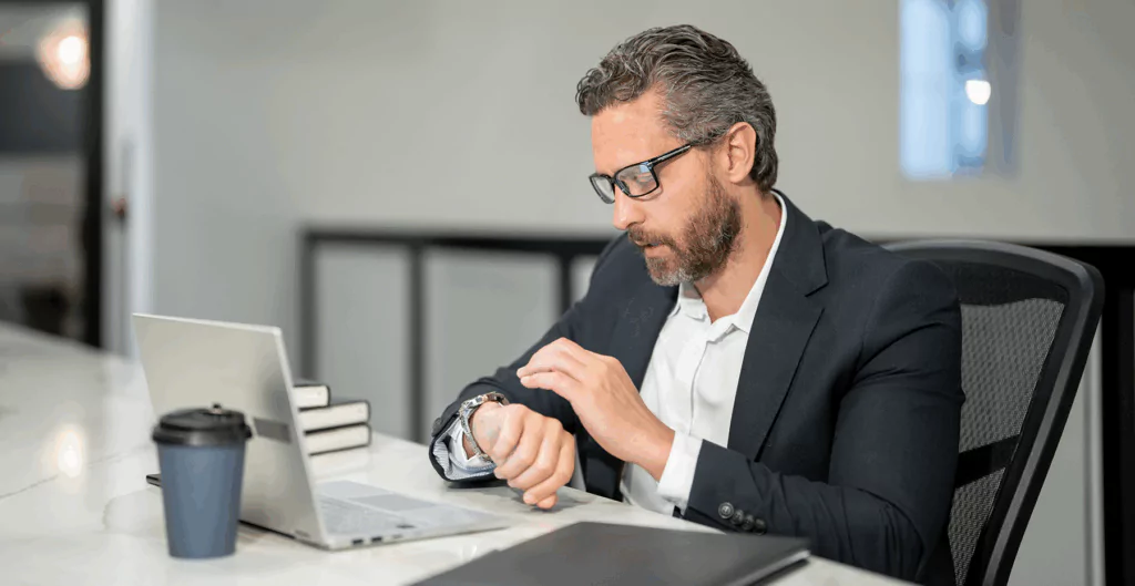 Homem de negócios em escritório verificando o relógio, usando blazer e óculos, com laptop e caderno na mesa