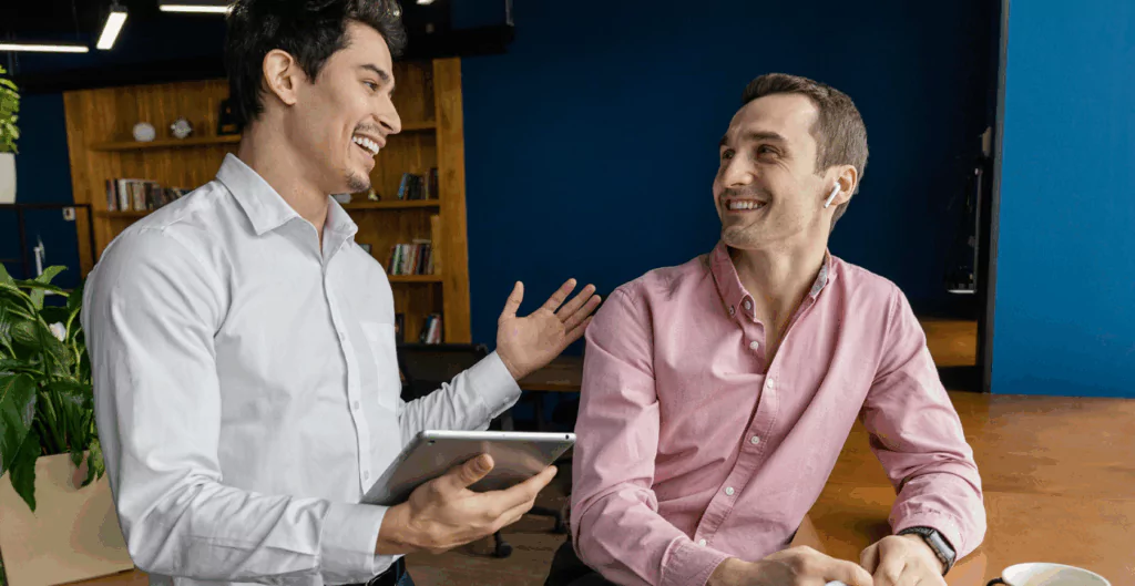 Homens profissionais conversando e sorrindo em ambiente de trabalho, ambiente de escritório moderno com estantes de livros ao fundo, promovendo interação e colaboração.