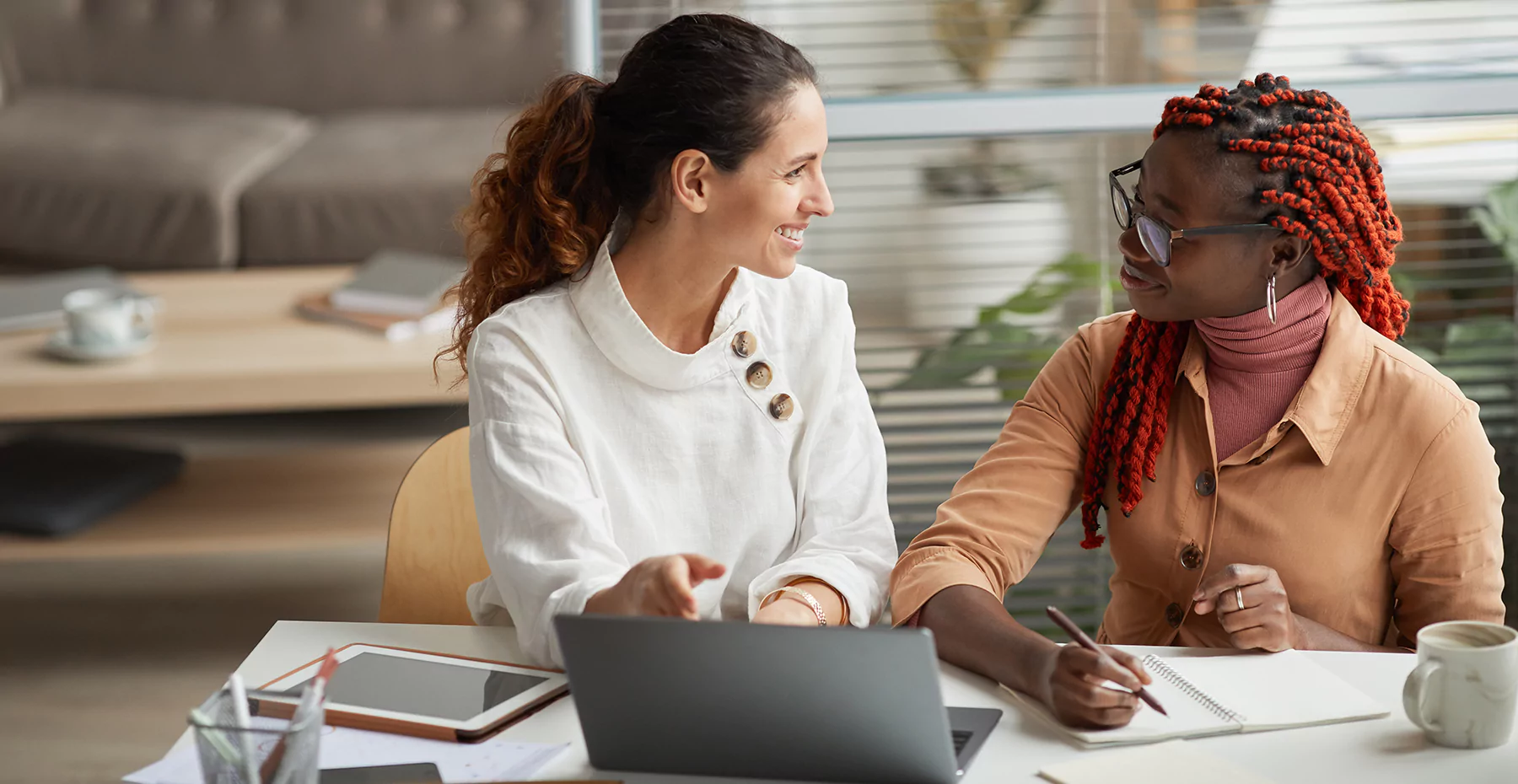 Duas mulheres conversando durante uma reunião de trabalho em um ambiente de escritório, com um laptop, blocos de anotação e uma caneca na mesa.