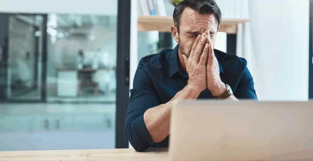 Homem sentado à mesa com laptop, cobrindo o rosto em sinal de exaustão, ilustrando disforia de carreira causada por sobrecarga, estresse mental e pressão no ambiente profissional.