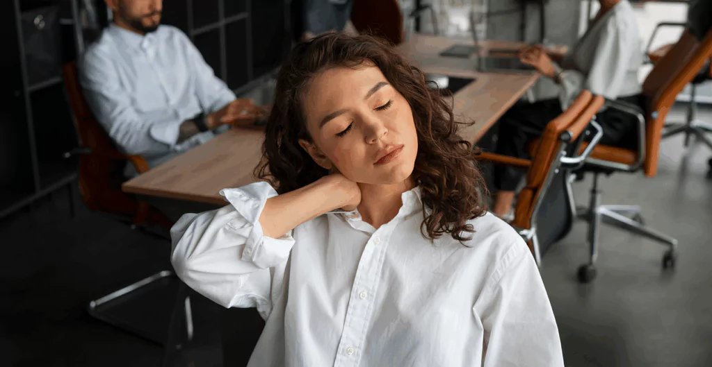 Mulher jovem demonstrando cansaço ou sonolência em ambiente de escritório ou reunião, com expressão de fadiga e olhos fechados, rodeada de colegas de trabalho.