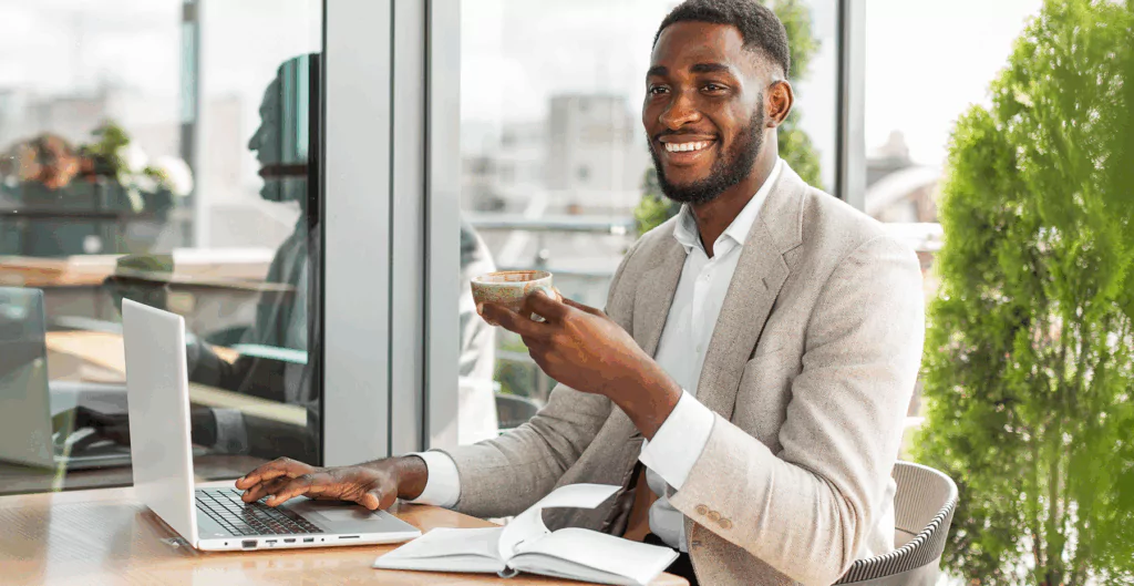 Homem usando laptop e celular em ambiente de escritório com muita luz natural e plantas verdes ao fundo.