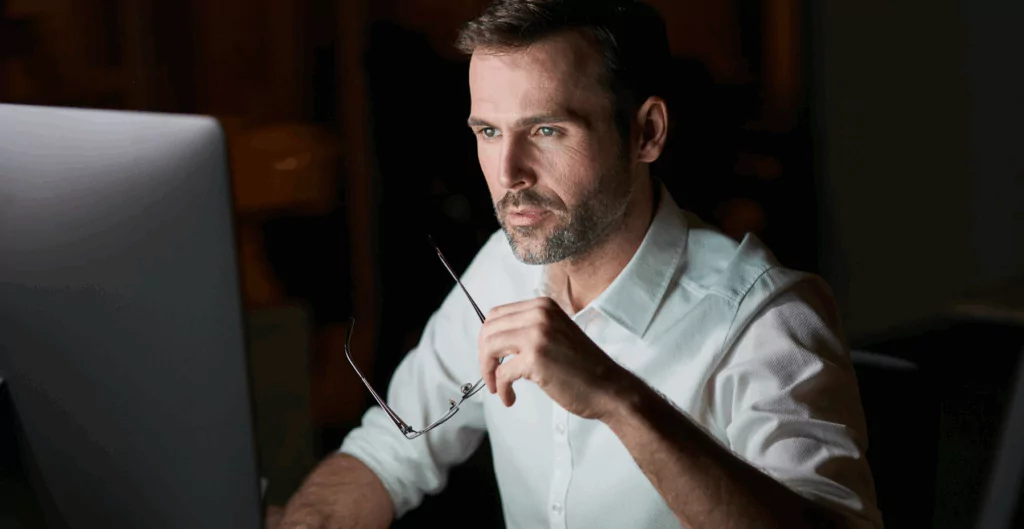 Homem senado, segurando um óculos e olhando a tela de um computador