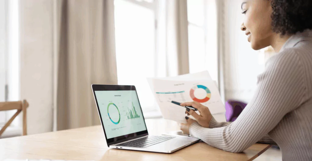 Mulher de cabelos cacheados analisando gráficos em uma reunião de negócios, com um laptop em uma mesa de escritório iluminada por luz natural.