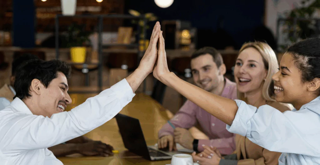 Quatro colegas de trabalho comemorando uma vitória ou conquistando uma meta juntos, com sorrisos e high five em ambiente de escritório moderna e acolhedora.