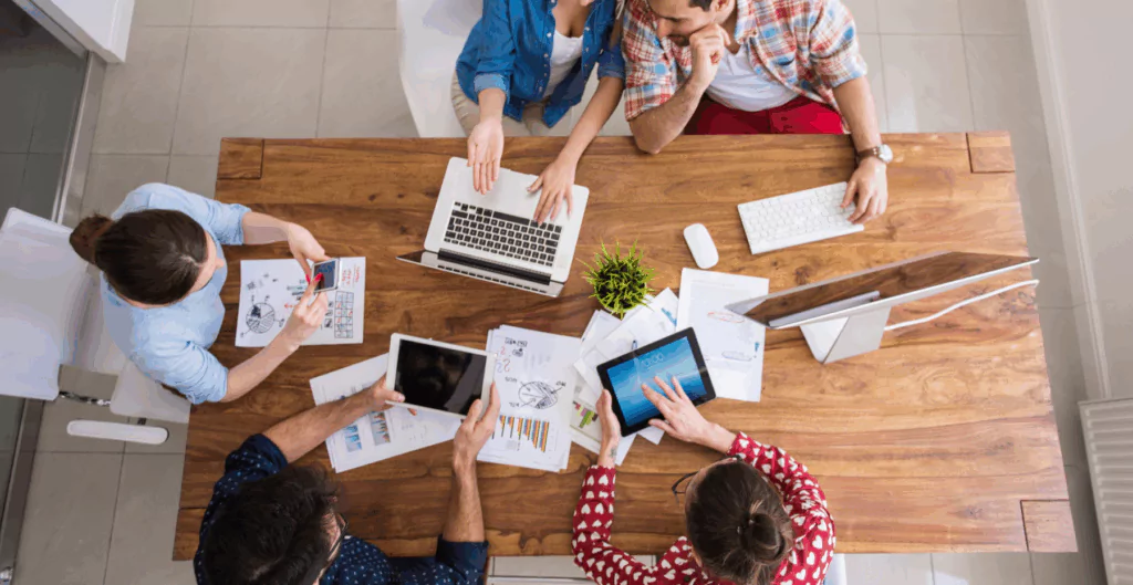 Equipe de trabalho colaborando em reunião de negócios, com laptops, tablets, gráficos e papéis sobre mesa de madeira. Ambiente moderno e organizado.