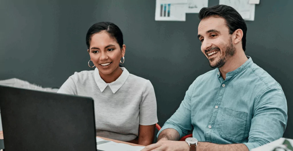 Duas pessoas sorridentes em uma reunião de negócios, discutindo ideias enquanto revisam documentos e utilizando um laptop em um ambiente profissional.