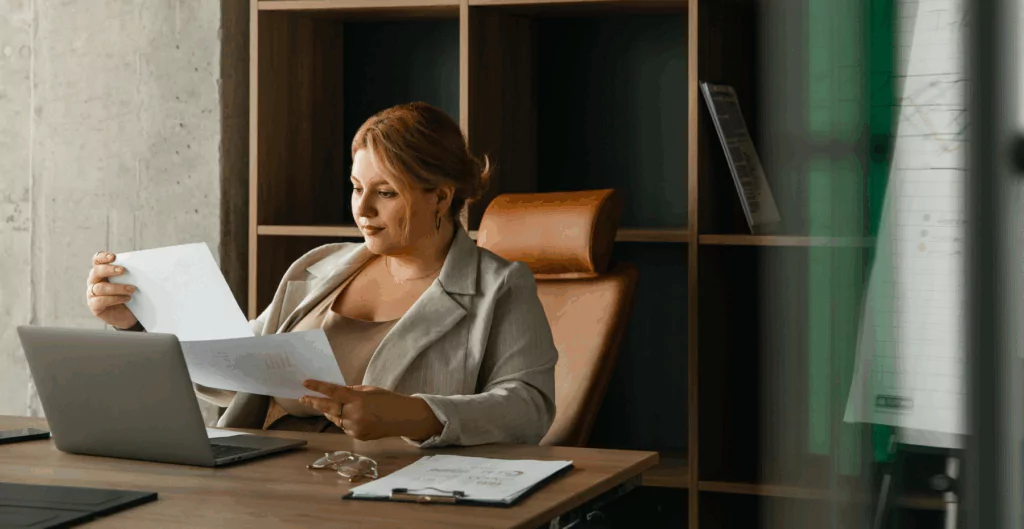 Mulher de negócios analisando documentos em escritório moderno, com computador portátil e escrita à mão, refletindo profissionalismo e organização.