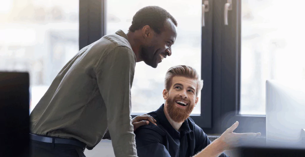 Homens profissionais sorrindo e conversando em ambiente de escritório moderno, demonstrando colaboração e comunicação eficaz.