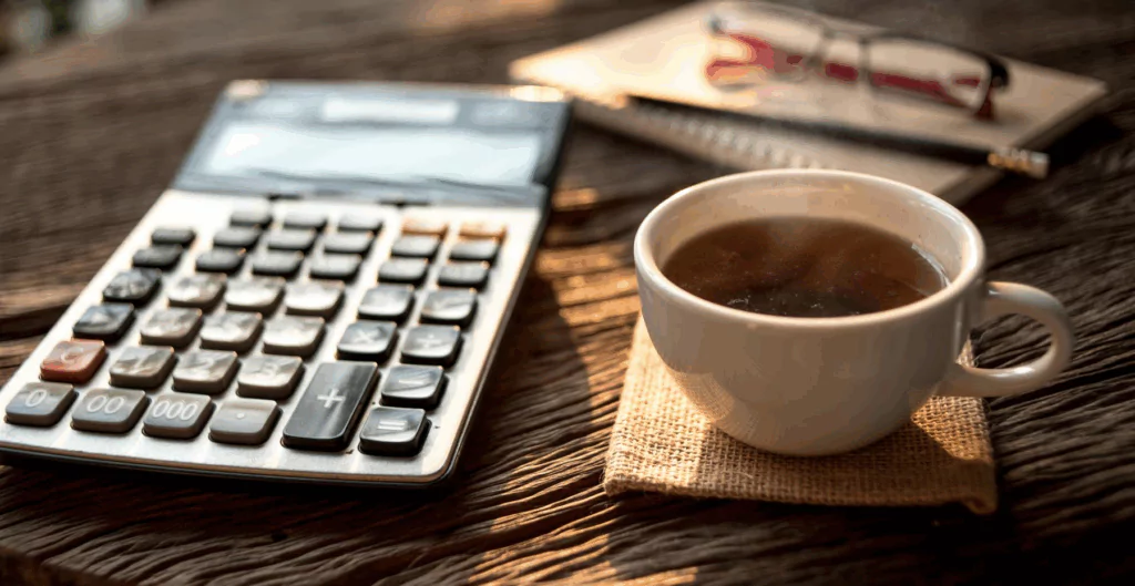 Calculadora, xícara de café e caneta sobre mesa de madeira, ambiente de trabalho caseiro ou escritório com iluminação natural.