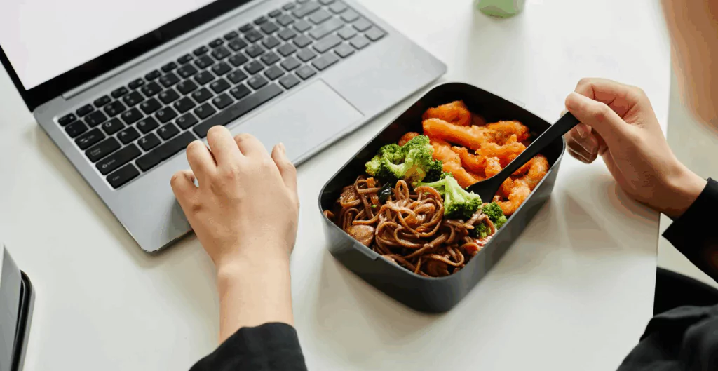 Pessoa usando um laptop enquanto come uma marmita com macarrão, brócolis e cenoura, ideal para o trabalho ou estudos.