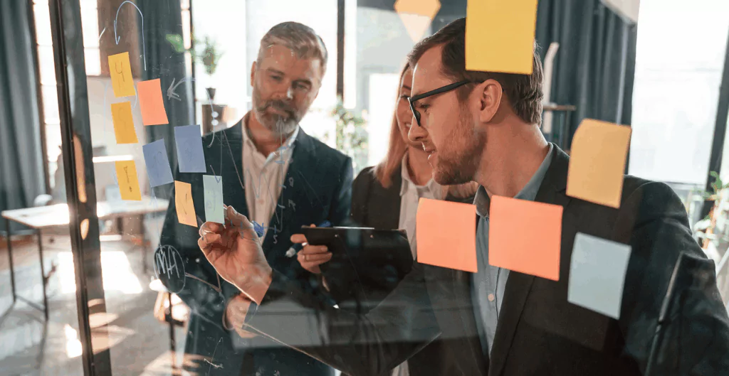 Equipe de profissionais de negócios planejando estratégias em reunião com uso de quadro de post-its e tecnologia em ambiente de escritório moderno.