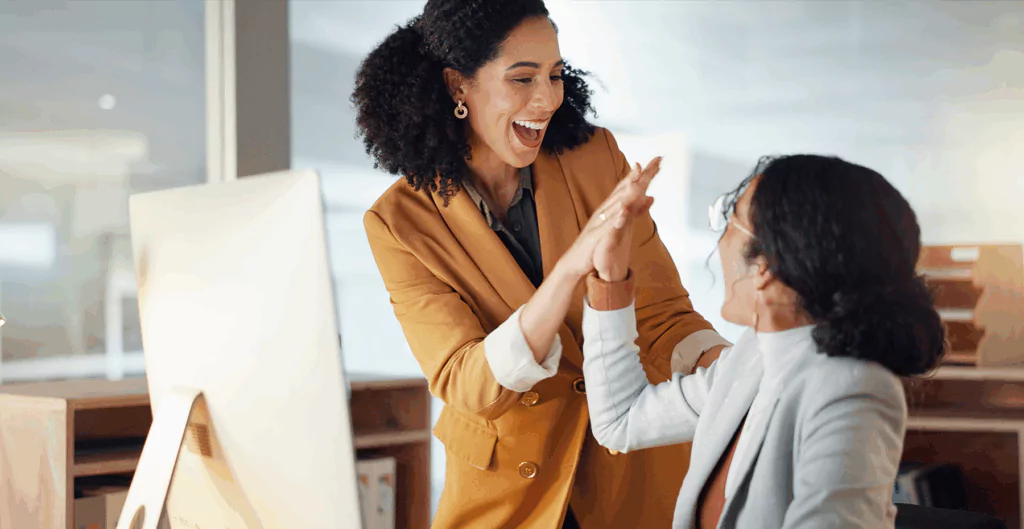 Duas mulheres de negócios comemorando uma vitória no ambiente de escritório, com expressões de felicidade e entusiasmo, destacando a colaboração profissional.