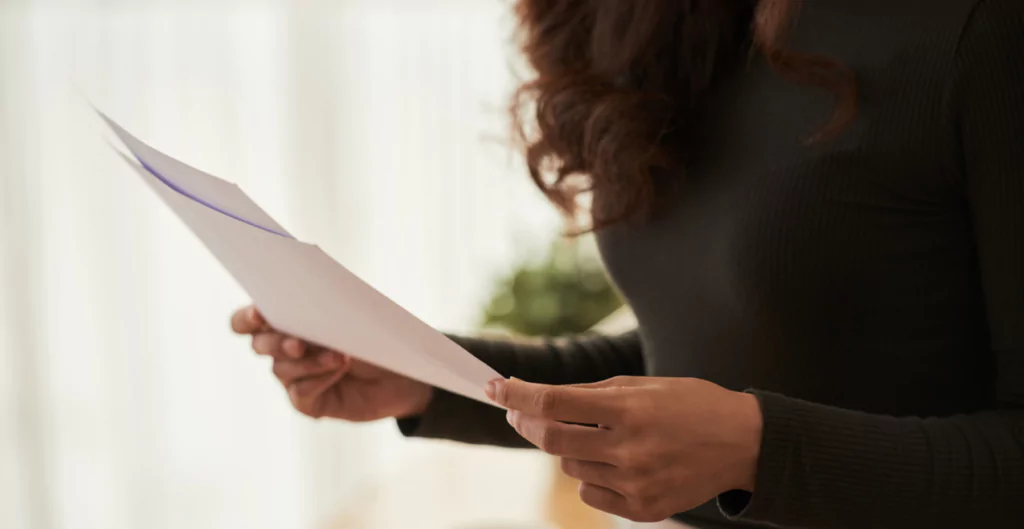 Mulher segurando uma folha de papel com foco nas mãos, evidenciando atividade de leitura ou revisão de documentos em ambiente interno bem iluminado.