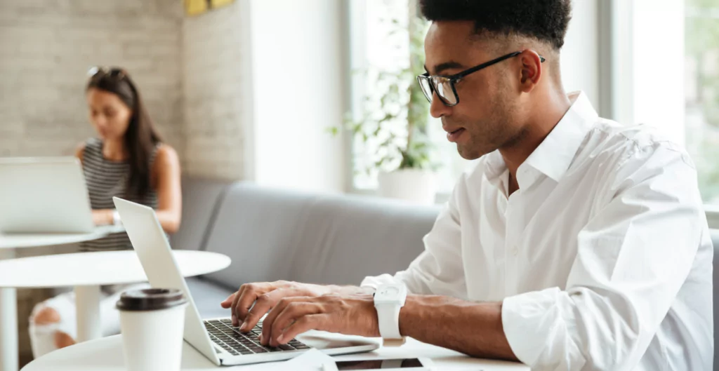 Homem afrodescendente usando laptop em ambiente de coworking com mulher ao fundo, conceito de trabalho remoto e tecnologia.