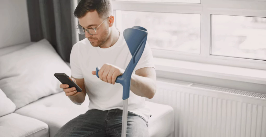 Homem usando um dispositivo de assistência para mobilidade em casa enquanto checa o celular. Ambiente bem iluminado com janelas grandes ao fundo.