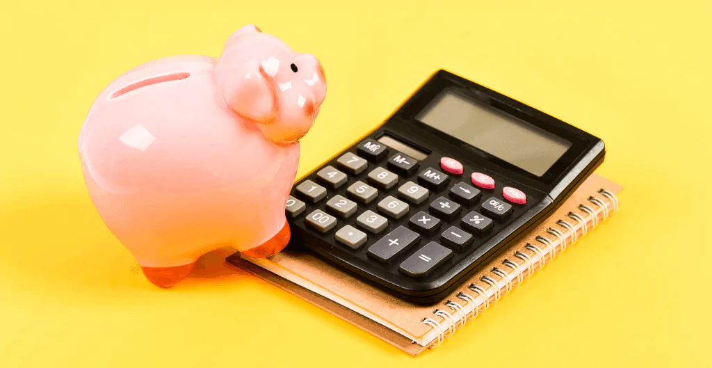 	
Porquinho de cerâmica rosa ao lado de uma calculadora solar e caderno em fundo amarelo, representando economia e finanças pessoais.