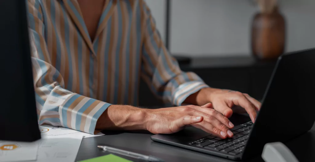 Pessoa com camisa listrada digitando em um notebook sobre mesa escura.