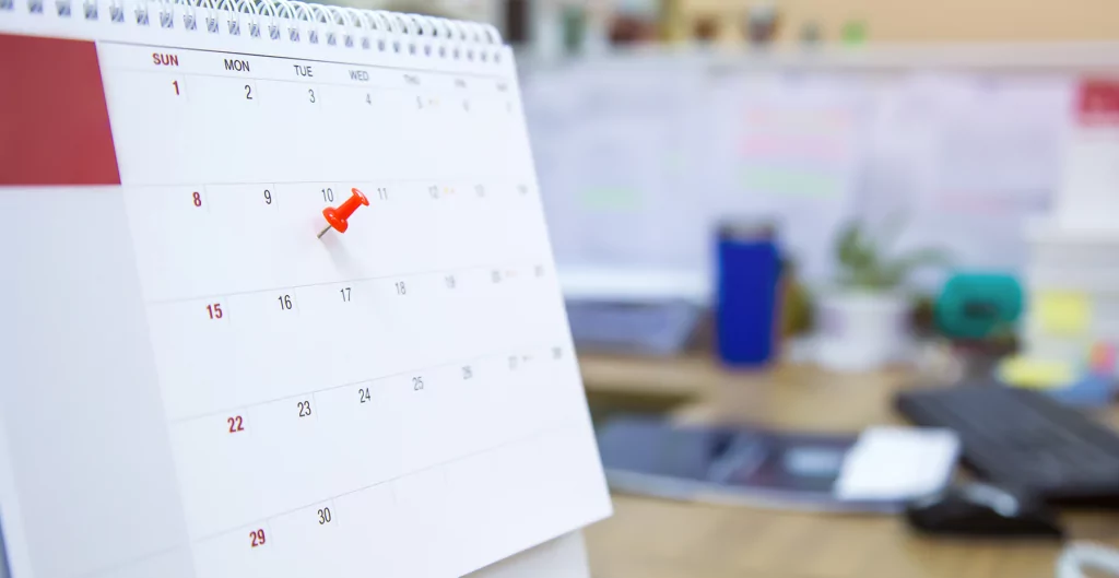 Calendário de mesa com um alfinete vermelho marcando o dia dez em um escritório desfocado ao fundo.