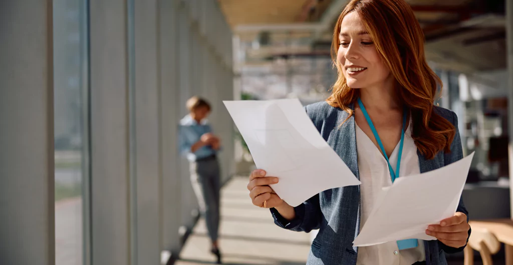 Mulher vestindo blazer cinza segura e analisa folhas de papel em um corredor de escritório iluminado.