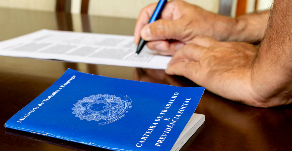 Pessoa assinando documento ao lado de uma Carteira de Trabalho e Previdência Social azul sobre a mesa.