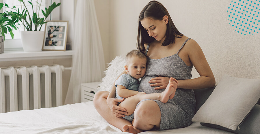 imagem de uma mulher sentada na cama dentro de um quarto segurando uma criança e com as mãos na barriga