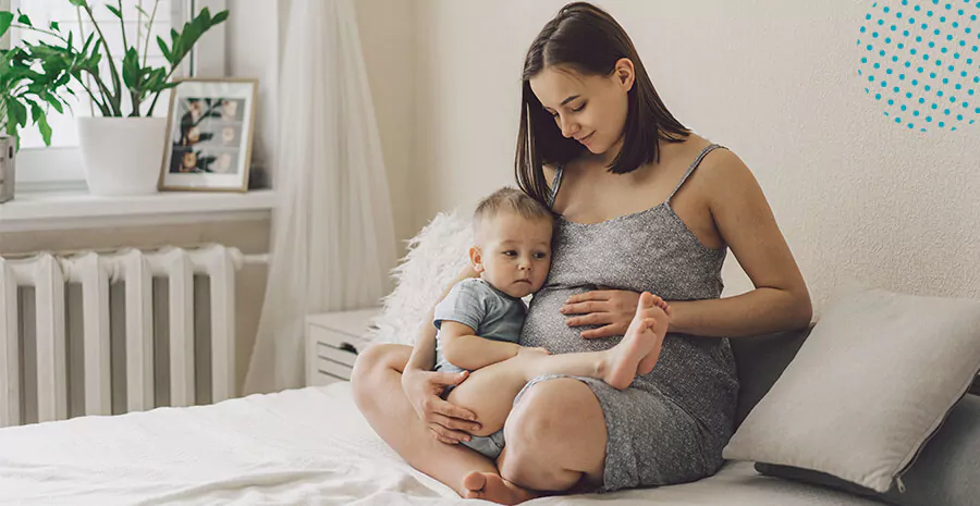 imagem de uma mulher sentada na cama dentro de um quarto segurando uma criança e com as mãos na barriga