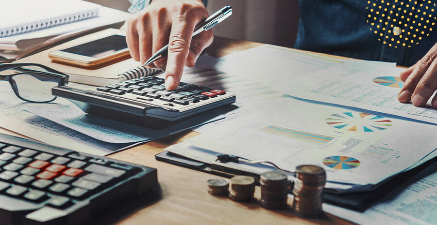 imagem de uma mesa de trabalho rodeada de papeis com uma pessoa utilizando uma calculadora