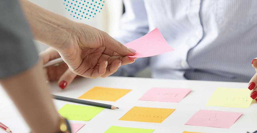 imagem de duas mãos femininas separas por uma mesa cheia de post-its amarelos, laranjas e rosas. Uma das mãos leva um post its rosa em direção a mesa