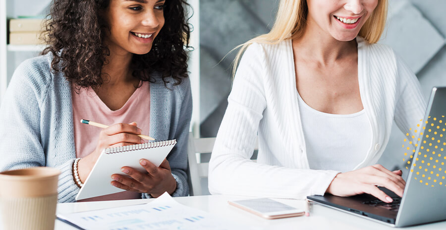 imagens de duas mulheres, uma morena e outra caucasiana, sentadas na mesa de escritório. Aparentam felicidade