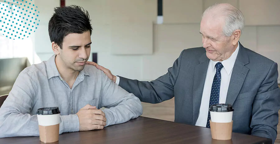 imagem de dois homens sentados, apoiando numa mesa com dois copos de café e um homem falando com o outro