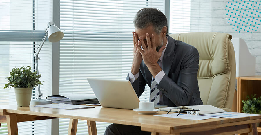 imagem de um homem sentado numa cadeira de escritório de frente para uma mesa com notebook e as mãos no rosto