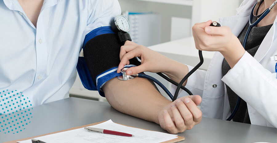 imagem de um médico examinando um paciente com medidor de pressão sob uma mesa com papel e caneta