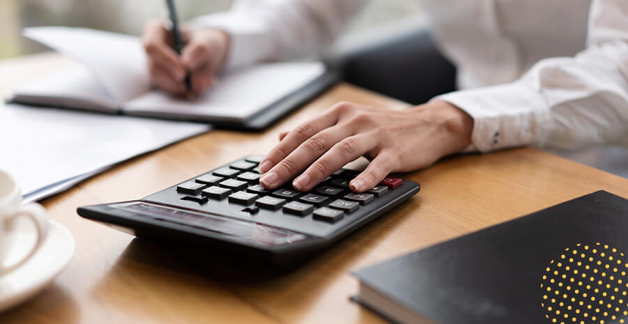 imagem de uma calculadora sob uma mesa com cadernos e uma pessoa escrevendo enquanto utiliza a calculadora para definir o salário mínimo