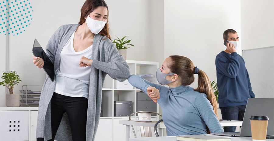 imagem de duas mulheres de máscaras no ambiente de trabalho se cumprimentando pelos cotovelos