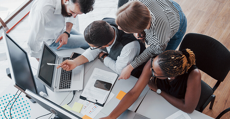 imagem de quatro pessoas diferentes trabalhando juntas, conversando e discutindo olhando para um computador sob uma mesa com papéis, celular e telas para o trabalho de outsourcing